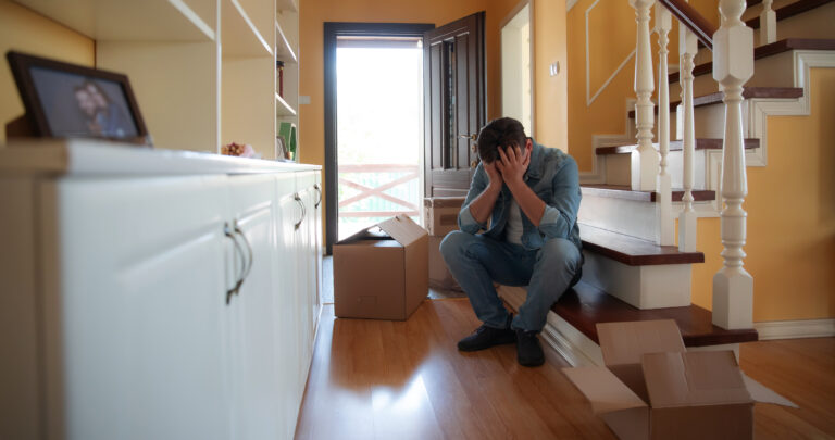 Man sits on the stairs with his head in his hands, overwhelmed by the pain of a recent divorce. Surrounded by moving boxes and a broken photo frame, he with deep emotional toll of separation